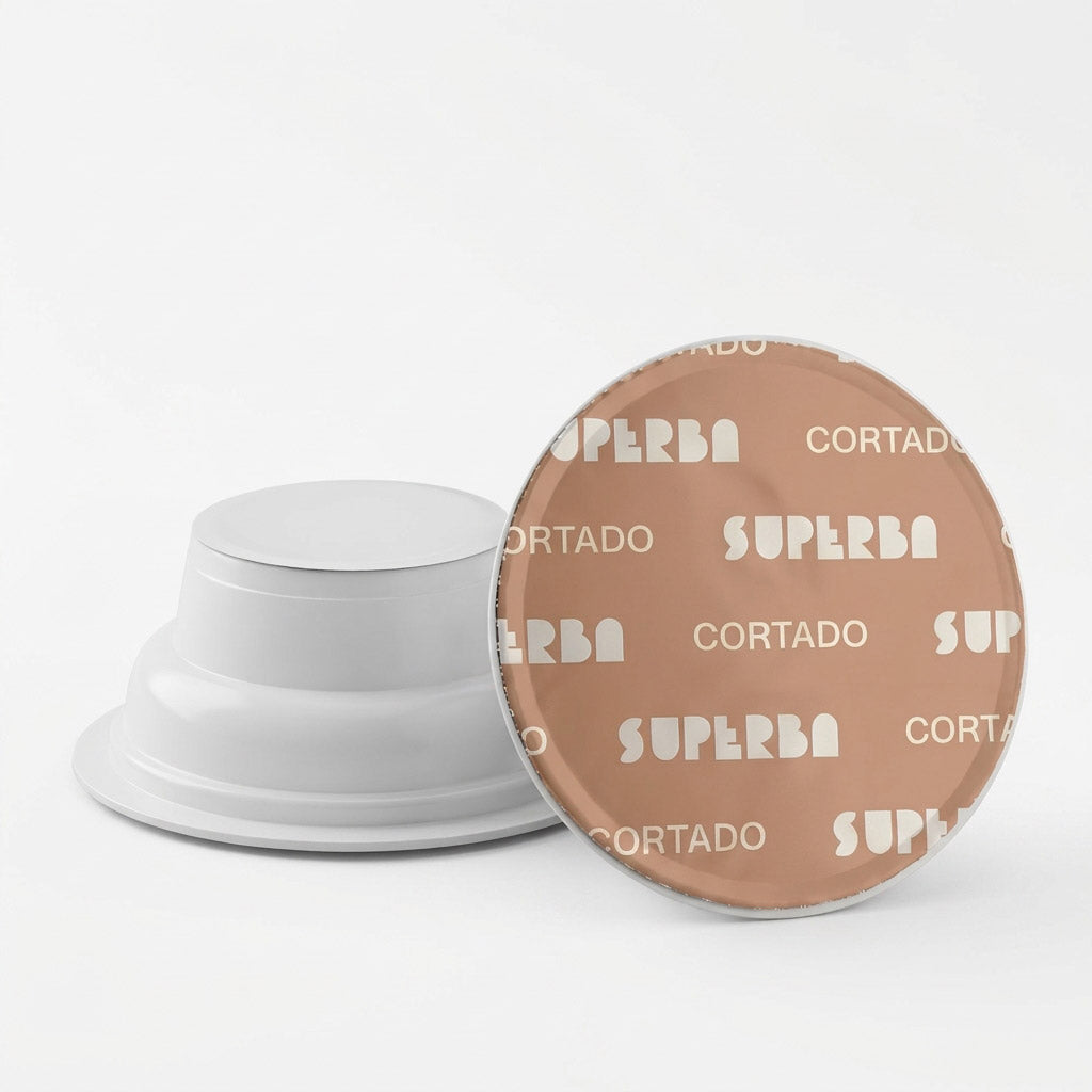 Dettaglio di due capsule Covim Superba Cortado: una mostrata di profilo con corpo bianco e l'altra vista dall'alto con pellicola beige chiaro e scritte bianche.